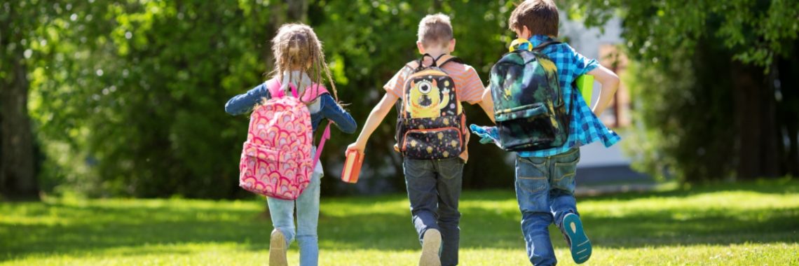 Three kids walking on green grass towards a set of trees in a park with unique and stylish backpacks on their backs.