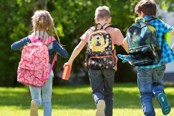 Three kids walking on green grass towards a set of trees in a park with unique and stylish backpacks on their backs.